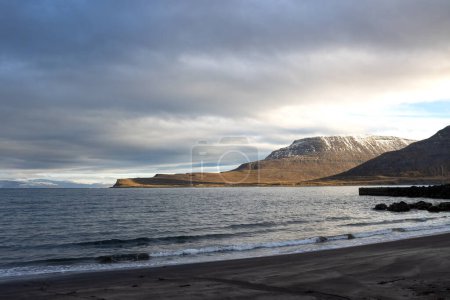 Dunkle vulkanische Küste des Atlantiks. Berge am Horizont, mit Schneegipfeln. Der Himmel ist stark bewölkt. Gebiet von Isafjordur, Westfjorde, Island.