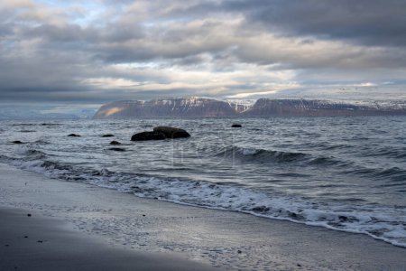 Foto de Costa volcánica oscura del océano Atlántico. Montañas en el horizonte, con picos de nieve. Cielo nublado intenso. Zona de Isafjordur, Westfjords, Islandia. - Imagen libre de derechos