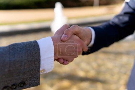 Photo for Anonymous businessmen in formal suits shaking hands while standing near blurred fountain during meeting - Royalty Free Image