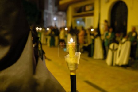 Candle held by Member of a brotherhood dressed in tunic and capirucho in an Holy Week procession at night. Devotee, copy space