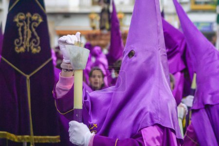 Photo for April 7th 2023 - Dona Mencia, Cordoba, Spain. Nazarene capirucho devotee fires candle in spanish Holly Week procession in windy day. Purple hood, white gloves, unrecognized person - Royalty Free Image