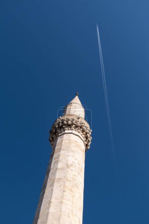 Mosque minaret against blue sky, flying plane leaving chemtrail