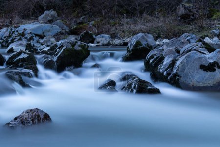 Gebirgsbach zur blauen Stunde mit seidigem Wasser, schneller Bachlauf um nasse Felsen in Langzeitbelichtung