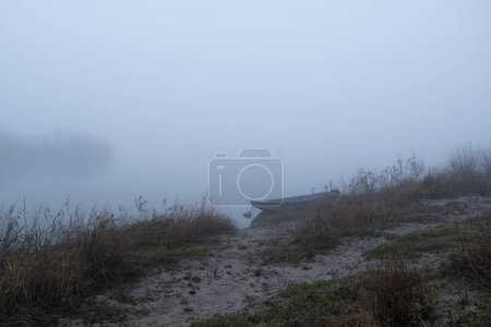 Schlammpfad führt zu festgemachtem Boot im Nebel, ruhiger nebliger Abend in Flussnähe, nautisches Schiff