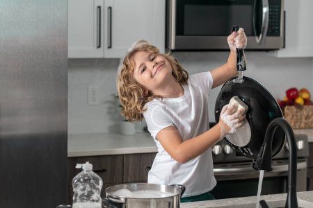 Foto de Divertido niño ayuda en el lavado de platos en la cocina. Lavavajillas botella líquida en fregadero de cocina y platos limpios - Imagen libre de derechos