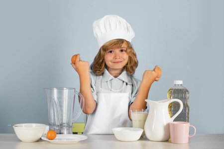 Foto de Retrato de un niño de 7, 8 años en gorra de cocinero y delantal sostienen los huevos haciendo ensalada de frutas y cocinando alimentos en la cocina. Linda pequeña rubia feliz chef sonriente - Imagen libre de derechos