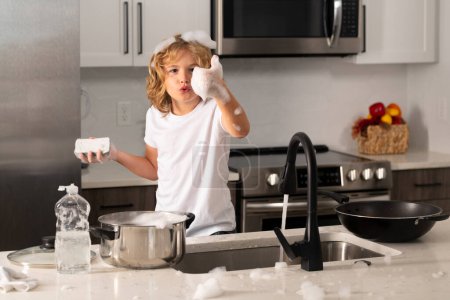 Foto de Niño lavando platos en el interior de la cocina. Limpie los platos lavados, lavavajillas líquido con espuma. Niño ayudando a sus padres con las tareas domésticas - Imagen libre de derechos