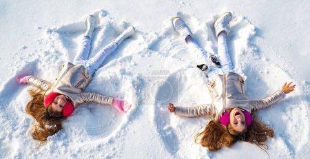 Téléchargez les photos : Des anges d'hiver. Les jeunes filles heureuses font des ailes d'ange de neige. Deux enfants font un ange de neige, couché sur la neige. Enfant excité jouant sur fond d'hiver. Jeux d'hiver avec neige pour les enfants - en image libre de droit
