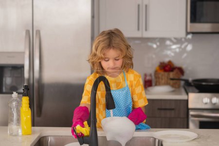 Foto de Niño lavar los platos en el interior de la cocina. Niño ayudando con las tareas domésticas. Concepto de limpieza doméstica y del hogar. Limpia los platos lavados, líquido lavavajillas con espuma - Imagen libre de derechos