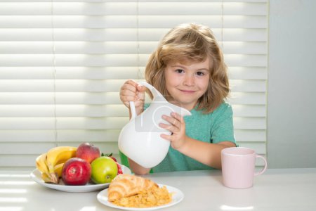 Foto de Fuuny pequeño niño vertiendo leche de vaca entera para el desayuno. Divertido niño rubio desayunando. Leche, verduras y frutas Alimentación saludable para niños - Imagen libre de derechos