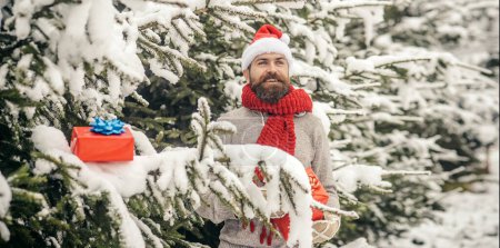 Foto de Hombre de Santa con caja de regalo al aire libre sobre fondo de nieve de invierno. Hombre en sombrero de santa con regalos de Navidad al aire libre. Fondo de invierno nevado. Santa al aire libre en nevadas llevando regalos - Imagen libre de derechos