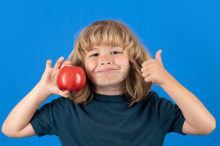 Foto de Un chico con pulgares arriba comiendo manzana. Retrato de niño lindo niño de pie aislado sobre fondo azul - Imagen libre de derechos
