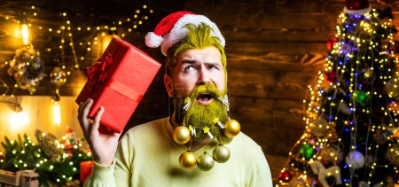 Téléchargez les photos : Père Noël avec un cadeau. Visage rapproché de jeune homme avec barbe décorée et chapeau de Père Noël - en image libre de droit