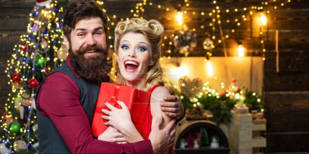 Photo for Beautiful Christmas couple smiling with gift. Retro woman with blonde hair and vintage bearded man near the Christmas tree. Couple in Christmas clothing hold gift. Gift wife emotions - Royalty Free Image