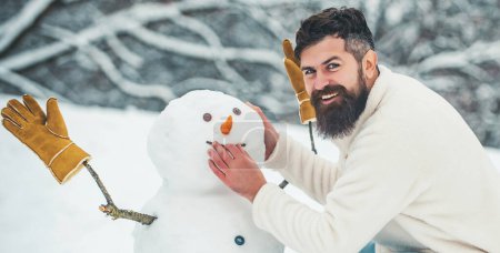 Téléchargez les photos : Des bonhommes de neige drôles. Un homme souriant heureux fait bonhomme de neige le jour ensoleillé de l'hiver. Homme faisant bonhomme de neige en plein air - en image libre de droit