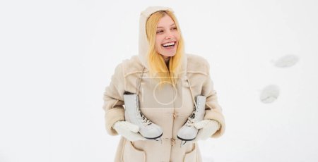 Foto de Mujer rubia joven en suéter y sombrero divertido sosteniendo patines blancos sobre el hombro en el frío día de invierno. Hermosa mujer caminando en el parque de invierno. Mujer en invierno nieve fondo - Imagen libre de derechos