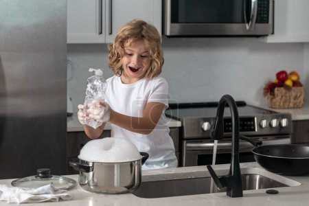Foto de Un niño lindo lavando platos cerca del fregadero en la cocina - Imagen libre de derechos
