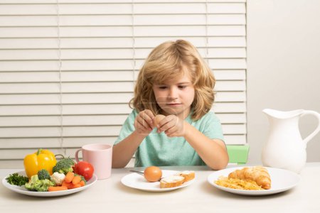 Foto de Niño comiendo huevo. Divertido niño rubio desayunando. Leche, verduras y frutas Alimentación saludable para niños - Imagen libre de derechos