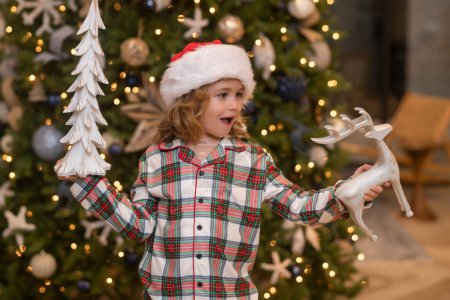 Téléchargez les photos : Des jouets de Noël. Un enfant incroyable tient une balle de Noël. Le garçon se réjouit de la fête, Noël, Nouvel An. Le gamin se tient contre le sapin de Noël - en image libre de droit