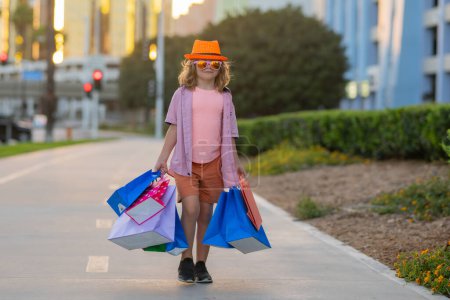 Photo for Cute little boy in summer fashion clothes goes shopping. Happy child with shopping packages in hands. Shopper child with shopping bag outdoor. Cute kid walking on street carrying shopping bags - Royalty Free Image