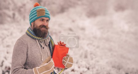 Téléchargez les photos : Portrait d'homme tenant boîte cadeau à l'extérieur au pôle Nord. Homme heureux avec cadeaux de Noël en plein air. Fond hivernal enneigé. Photo de Père Noël heureux en plein air dans les chutes de neige portant des cadeaux - en image libre de droit