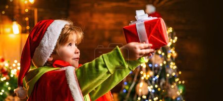 Téléchargez les photos : Enfant garçon en chapeau de Noël Santa détient une boîte-cadeau et fond décoré à la maison. Joyeux Noël et bonne année. Petit garçon avec des cadeaux de Noël - en image libre de droit