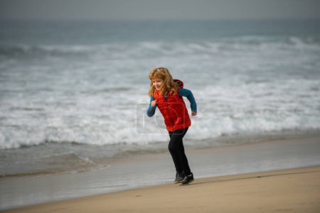 Téléchargez les photos : Enfant traversant l'eau près du rivage le long de la plage de la mer. Un garçon court le long de la côte. Enfant coureur joggeur courir dans la nature. Mode de vie sain et actif des enfants - en image libre de droit