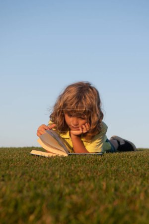 Téléchargez les photos : École de plein air, les enfants motivation apprentissage. Un enfant lit un livre sur le parc du printemps. Détente sur l'herbe dans le jardin. Éducation en plein air - en image libre de droit