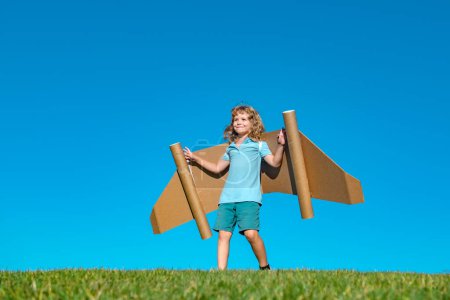 Téléchargez les photos : Child boy plays in an astronaut dreams of space. Happy child play with toy plane cardboard wings against blue sky. Kid having fun in summer field outdoor. Portrait of boy with paper wings - en image libre de droit