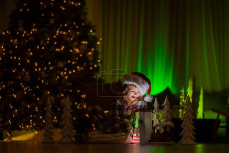 Téléchargez les photos : Cadeaux d'ouverture d'enfant la veille de Noël. Enfant garçon 7-8 ans en pyjama de Noël profitant d'une soirée de vacances d'hiver à la maison près de l'arbre de Noël de nuit - en image libre de droit