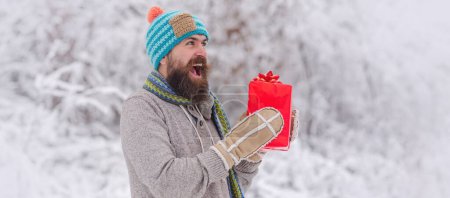 Foto de Retrato de hombre excitado en sombrero de punto y suéter de invierno celebrar regalo. Hombre feliz con regalos de Navidad al aire libre. Fondo de invierno nevado. Foto de feliz Santa al aire libre en nevadas llevando regalos - Imagen libre de derechos