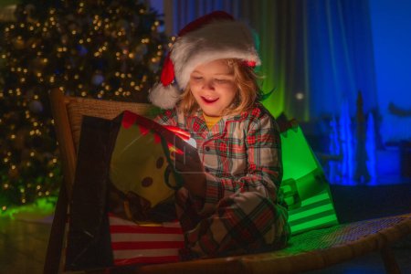 Photo pour Mignon sac cadeau d'ouverture d'enfant dans la chambre décorée pour la nuit de Noël. Enfant garçon 7-8 ans en pyjama de Noël profitant d'une soirée de vacances d'hiver à la maison près de l'arbre de Noël de nuit - image libre de droit