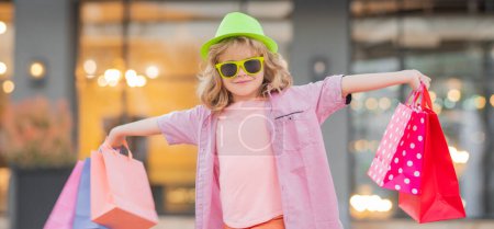 Photo for Funny fashion child model. Child on shopping. Portrait of a kid with shopping bags. Fashion child in shirt, sunglasses and hat. Shopping mall. Shopping on black friday - Royalty Free Image