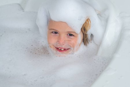 Foto de Cara de niños en espuma. Niño divirtiéndose en el baño con burbujas. Niño feliz disfrutando del baño. Niño sonriendo en el baño con espuma de jabón. El niño se baña en un baño con espuma - Imagen libre de derechos
