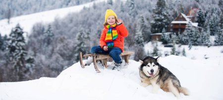 Photo for Boy with dog sledding on winter mountain, enjoying a sledge ride in a beautiful snowy winter nature. Winter fun kids activities - Royalty Free Image