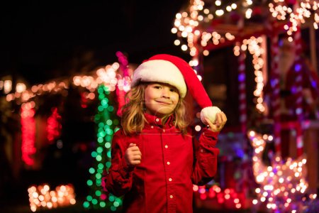 Téléchargez les photos : Enfant sur la décoration de Noël devant une maison de nuit. Soirée de Noël en arrière-plan de la maison de nuit avec guirlandes - en image libre de droit