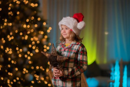 Téléchargez les photos : Enfant en santa chapeau sur le devant de l'arbre de Noël de nuit arrière-plan de la maison la veille de Noël. Le gamin s'amuse près du sapin de Noël la nuit à la maison - en image libre de droit
