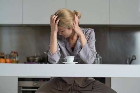 Téléchargez les photos : Femme déprimée dans la cuisine le matin. Femme bouleversée assise dans la cuisine regardant le téléphone le matin et bavardant smartphone. Triste femme au foyer fatiguée - en image libre de droit