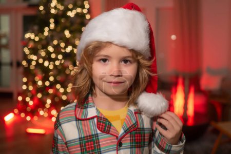 Téléchargez les photos : Enfant en santa chapeau sur le devant de l'arbre de Noël de nuit arrière-plan de la maison la veille de Noël. Joyeux enfant en pyjama de Noël à carreaux profitant d'une soirée de vacances à la maison - en image libre de droit