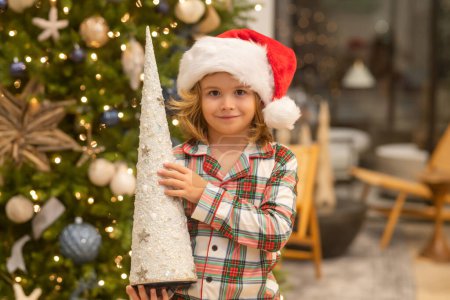 Téléchargez les photos : Joyeux enfant drôle dans le chapeau de Père Noël près de l'arbre de Noël. Noel et Nouvel An concept - en image libre de droit