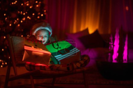 Photo for Child open present gift with magic light. Lighting present gift bag. Kid in santa hat on front of night Christmas tree home background on Christmas Eve - Royalty Free Image