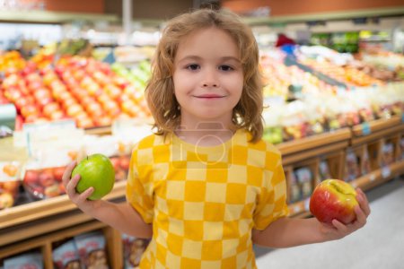 Téléchargez les photos : Un enfant tient des pommes à l'épicerie. Le gamin choisit des légumes et des fruits frais dans le magasin. Enfant achetant de la nourriture dans un supermarché. J'achète à l'épicerie. Epicerie au supermarché - en image libre de droit