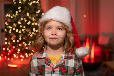 Téléchargez les photos : Enfant en chapeau de Père Noël sur le devant de l'arbre de Noël de nuit arrière-plan de la maison la veille de Noël - en image libre de droit