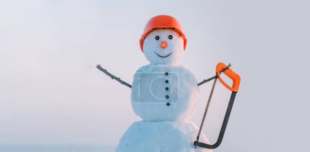 Téléchargez les photos : Snowman constructeur dans la construction casque tenir scie. Bonhomme de neige en casque sur le fond extérieur de la neige. Bannière de Noël avec bonhomme de neige. Bonhomme de neige prêt pour les travaux de construction et de réparation - en image libre de droit