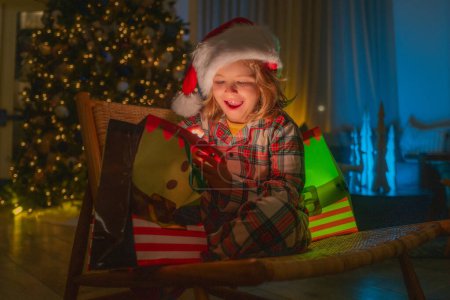 Téléchargez les photos : Mignon enfant ouvre boîte cadeau dans la chambre décorée pour la nuit de Noël. Enfant en pyjama de Noël profitant soirée de vacances d'hiver à la maison près de l'arbre de Noël de nuit - en image libre de droit