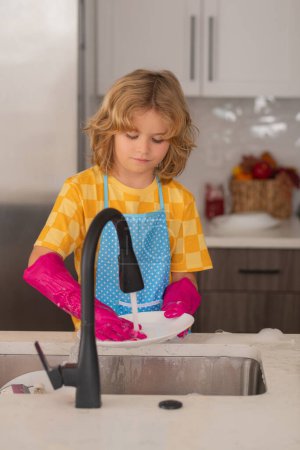 Téléchargez les photos : Nettoyage à la maison. Lave-vaisselle pour enfants dans la cuisine intérieure. Un enfant qui aide au ménage. Nettoyage et entretien ménager concept. Poussière et gants pour enfants pour le nettoyage - en image libre de droit