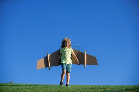Téléchargez les photos : Enfant drôle jouant avec des ailes d'avion à l'extérieur. Joli garçon jouant au pilote et rêvant de devenir pilote d'aviateur. Le gamin s'amuse avec des ailes en papier dehors. Vacances d'été et concept de voyage pour enfants - en image libre de droit