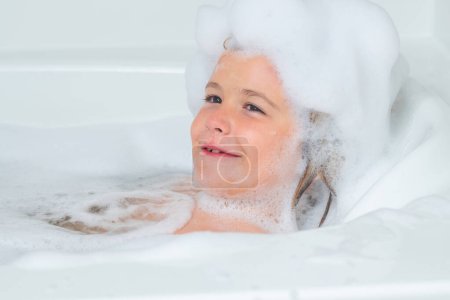 Foto de Champú para niños. Niño lavándose con una burbuja en el baño. Lindo niño se baña, acostado en un baño blanco con espuma de jabón y champú - Imagen libre de derechos