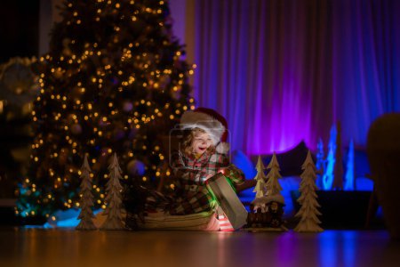 Photo for Child opening presents on Xmas eve. Happy little kids in matching red and green striped pajamas near Christmas tree in living room with traditional Christmas tree - Royalty Free Image