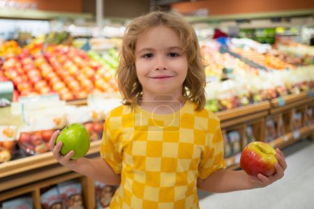 Téléchargez les photos : Un enfant tient des pommes à l'épicerie. Portrait d'enfant dans un magasin d'alimentation ou un supermarché. Un gamin qui va faire du shopping. Aliments sains pour enfants - en image libre de droit